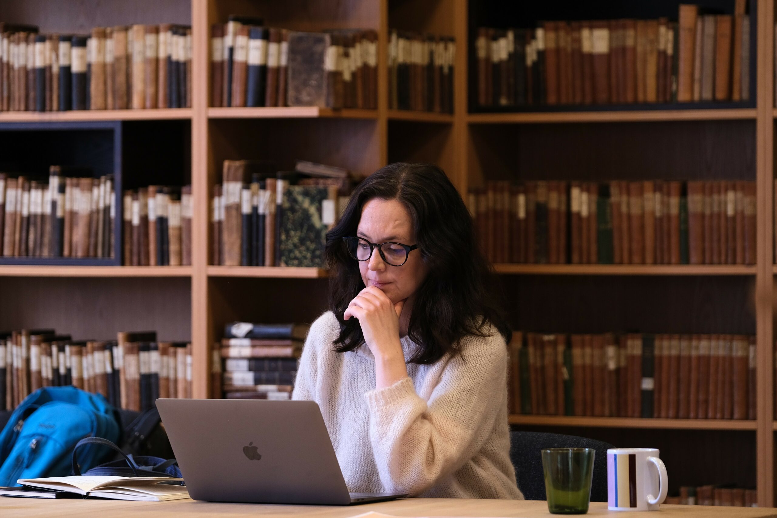 En kvinne i briller og en kremgenser arbeider ved en bærbar datamaskin ved et bibliotekbord med hyller fulle av bøker i bakgrunnen.
