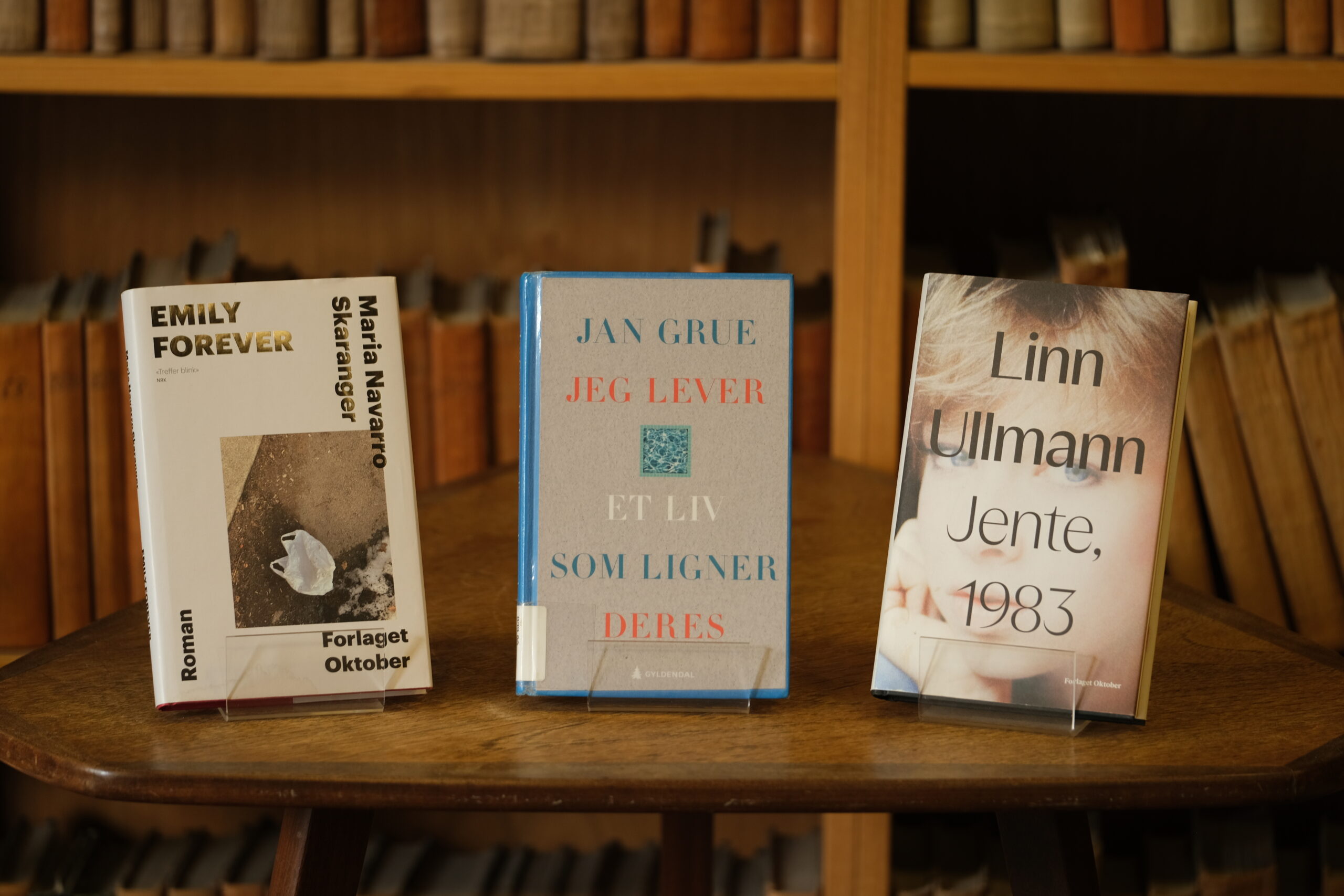 Tre bøker utstilt på en trehylle med titler av Maria Navarro Skaranger, Jan Grue og Linn Ullmann på norsk, med uskarp bakgrunn av bokhyller.