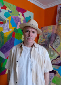 Mann iført stråhatt og hvit skjorte står foran fargerike abstrakte malerier i et rom med oransje vegger.