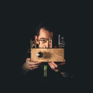 En mann som holder et særegent trekamera i vintagestil med messingkomponenter, med et fokusert uttrykk, mot en mørk bakgrunn.