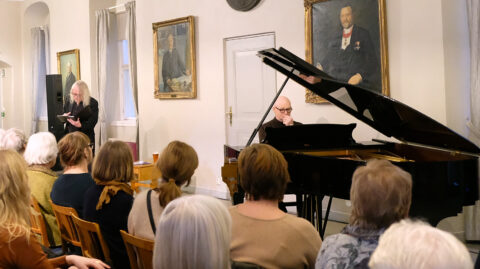 En pianist opptrer ved et flygel mens en kvinne står og leser for et oppmerksomt publikum i et rom utsmykket med portretter.