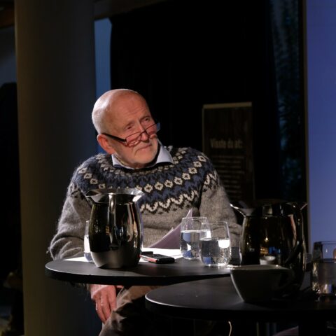 Eldre mann med briller og en mønstret genser som sitter ved et bord med en karaffel og vannglass, og virker kontemplativ.