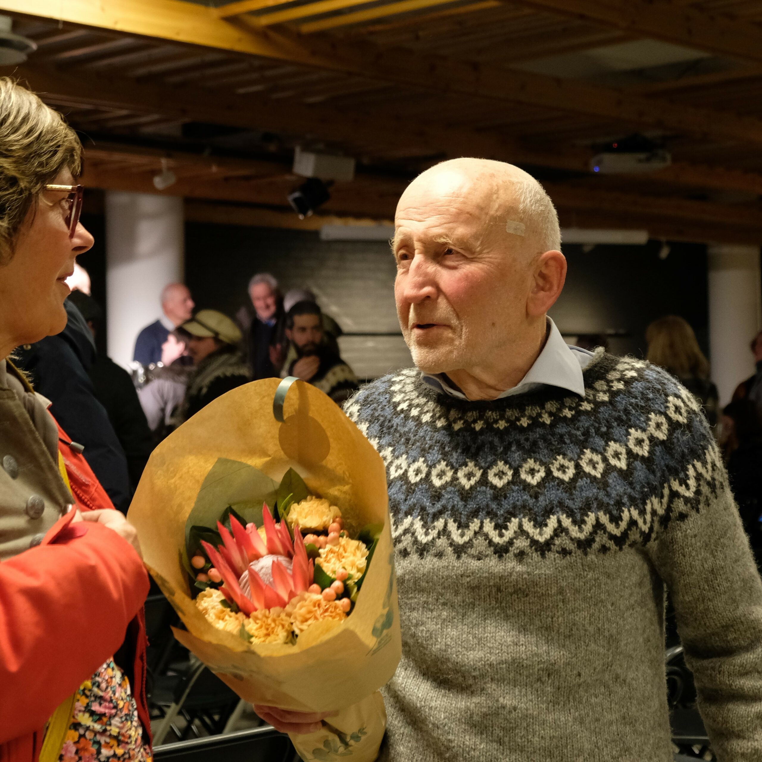 En eldre mann i en mønstret genser snakker med en kvinne mens han holder en blomsterbukett.