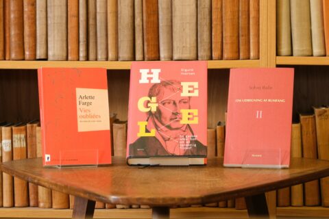 Tre bøker utstilt på et trebord mot et bakteppe av hyller fylt med gamle lærinnbundne bøker.