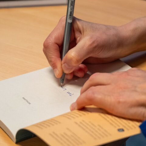 Et nærbilde av en persons hender som holder en oransje bok og signerer det hvite omslaget med en sølvpenn. Overflaten er laget av lyst tre.