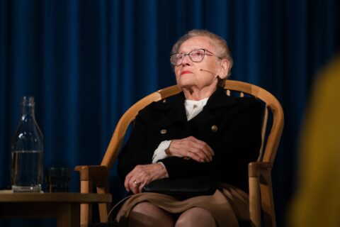 En eldre kvinne med briller og svart jakke sitter på en trestol med hendene foldet på fanget, foran en mørkeblå gardin. En glassflaske og et glass står på et bord ved siden av henne.