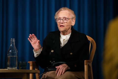 Eldre person med briller snakker mens han sitter på en stol, med blå gardinbakgrunn. En glassflaske og kopp står på et lite bord ved siden av dem.