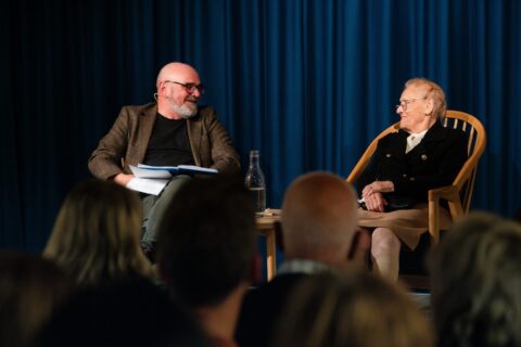 En eldre kvinne og en mann sitter overfor hverandre på scenen, engasjert i samtale foran et sittende publikum. Blå gardiner danner bakgrunnen.