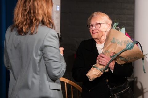 En eldre kvinne som holder en blomsterbukett står vendt mot en annen kvinne i en grå blazer som holder en mikrofon.