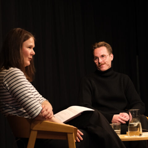 DSCF9548, Foto: Kristian Lillenes/Litteraturhuset i Trondheim To personer satt på scenen, engasjert i samtale. Den ene holder en notatbok, mens den andre smiler. En mugge og et glass vann står på bordet mellom dem.