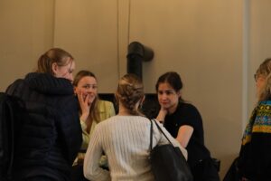 Fem kvinner står i en liten gruppe innendørs, engasjert i samtale med nøytrale og oppmerksomme uttrykk.