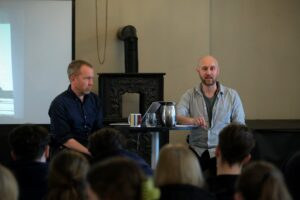 To menn sitter ved et bord foran et publikum og snakker på et innendørsarrangement med en tekanne, et krus og en skjerm i bakgrunnen.