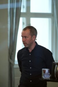 En mann med headset og mikrofon står innendørs foran et vindu med et glass vann og en metallmugge på et bord i nærheten.