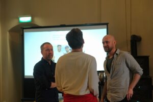 Tre menn står og snakker foran en projisert presentasjonsskjerm i et svakt opplyst rom.