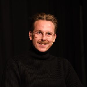 En mann med lysebrunt hår, briller og bart har på seg en svart polotrøye og sitter foran en mørk bakgrunn og smiler litt.