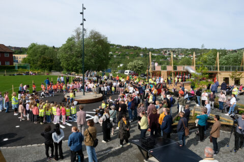 Tempe_overblikk, Foto: Kristian Lillenes/Litteraturhuset i Trondheim En stor gruppe mennesker, inkludert barn og voksne, samles på en utendørs lekeplass og parkområde på en solrik dag.