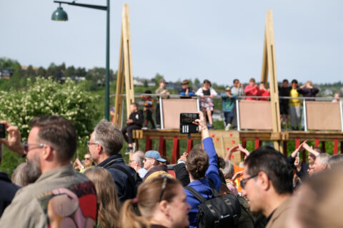 Tempe_publikum, Foto: Kristian Lillenes/Litteraturhuset i Trondheim En folkemengde, noen holder opp telefoner, samles utendørs nær en trebro på en solskinnsdag.