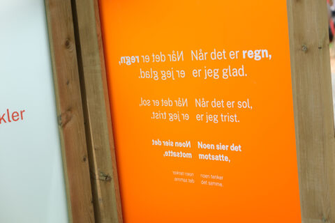 Tempe_regn, Foto: Kristian Lillenes/Litteraturhuset i Trondheim Tekst på en oransje og treaktig skjerm på norsk, som diskuterer følelser knyttet til regn og sol, med speilvendt og oppreist hvit tekst.